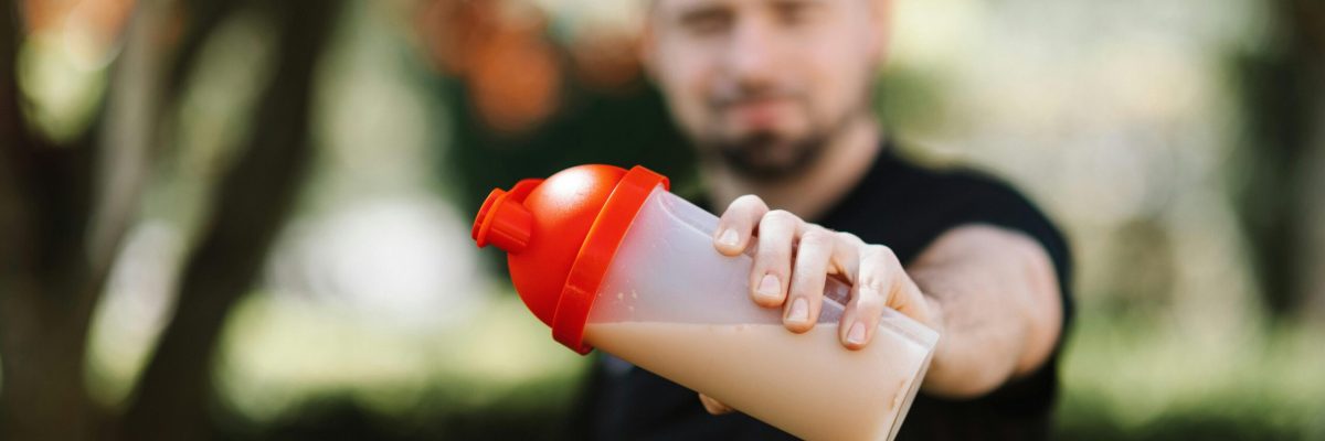 pexels-photo-4378525-4378525 Man presenting a protein shake in a plastic tumbler against a blurred nature background.